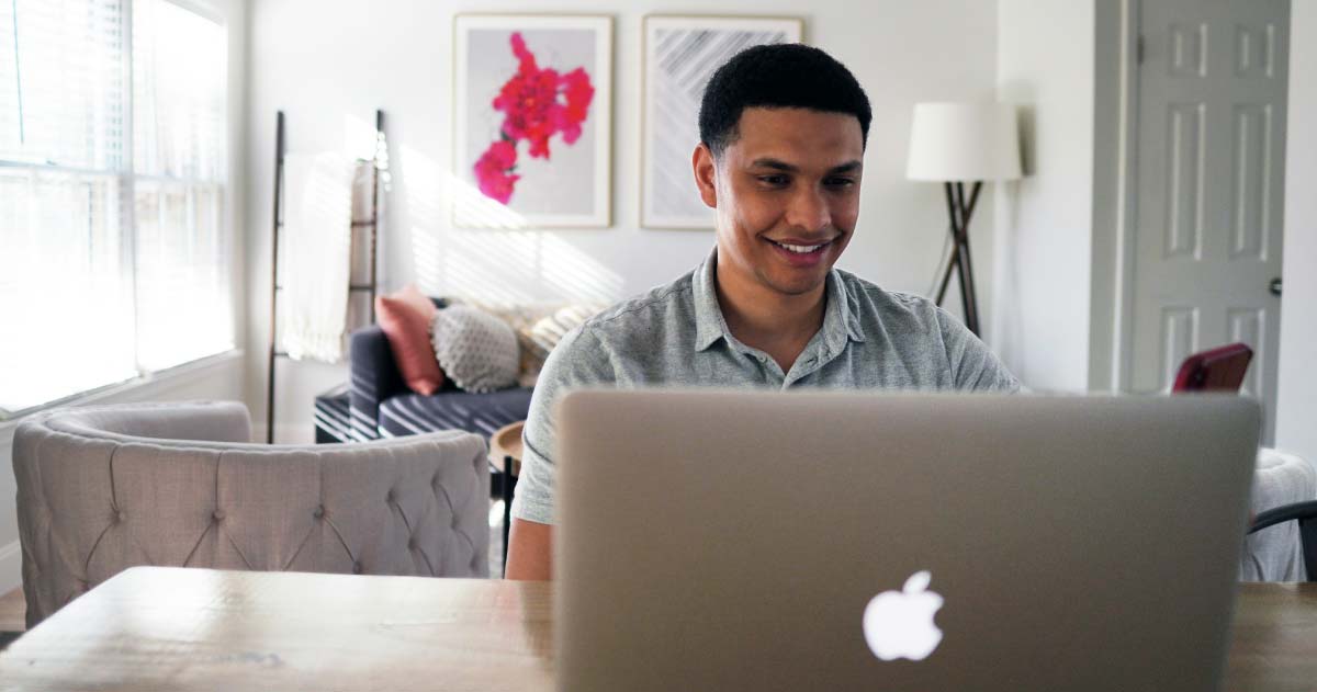 A person is sitting in a home at a desk with a laptop and smiling